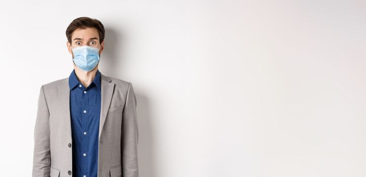 Health, pandemic and business concept. Surprised guy in suit and medical mask raising eyebrows, stare shocked at camera, white background.