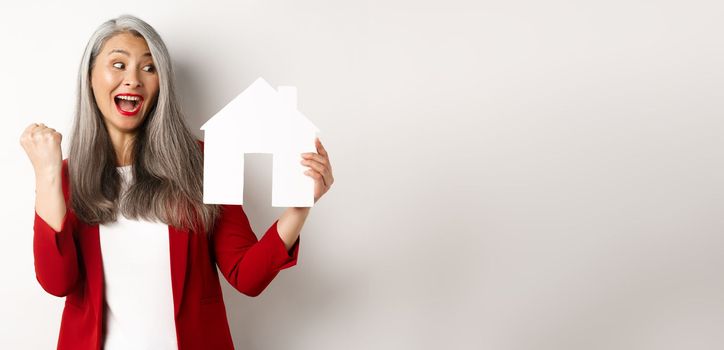 Cheerful asian senior woman buying house, scream of joy and making fist pump while showing paper house cutout, standing over white background.