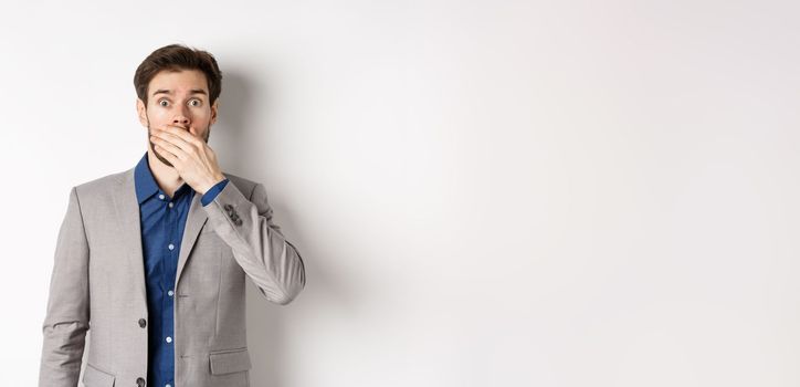 Shocked boyfriend in suit gasping, covering mouth with hand and pop eyes at camera startled, standing on white background.