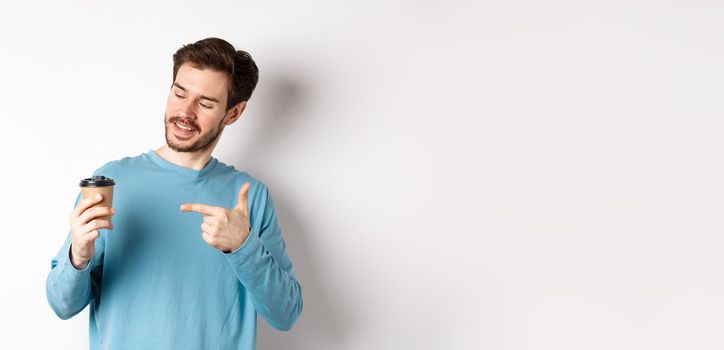 Handsome caucasian man pointing finger at coffee cup, recommending cafe, standing on white background satisfied.