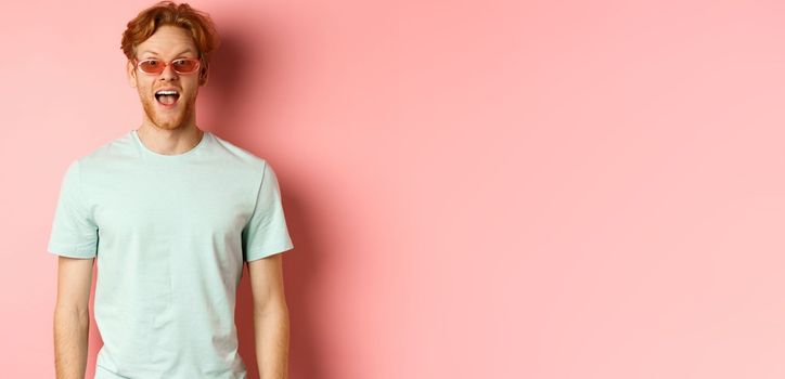 Image of surprised redhead man on vacation, wearing sunglasses with summer t-shirt, open mouth and saying wow amazed, standing over pink background.
