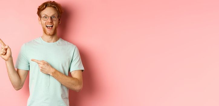 Portrait of cheerful young man with red hair, wearing glasses, pointing fingers at upper left corner and smiling, standing over pink background.