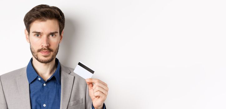 Finance. Close up of handsome bearded businessman in suit showing plastic credit card, standing serious on white background.