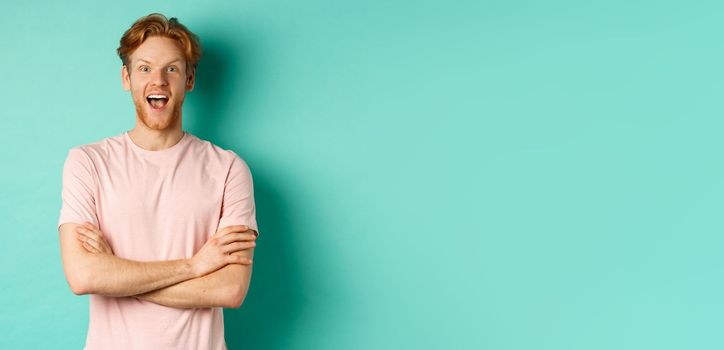 Enthusiastic redhead man in t-shirt checking out interesting promo, cross arms on chest and looking at camera with awe, standing over turquoise background.