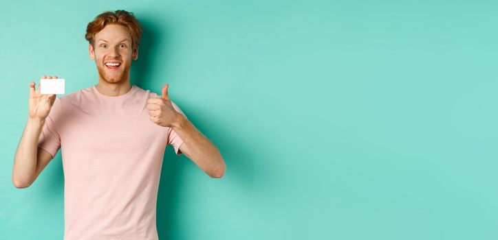 Cheerful male bank client in t-shirt showing thumb up and plastic credit card, smiling satisfied at camera, standing over turquoise background.