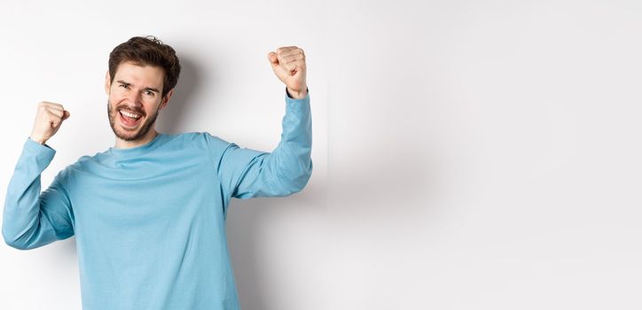 Image of happy young man triumphing, feeling like champion, winning and screaming in cheer, standing over white background.