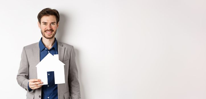 Real estate. Handsome young businessman buying property, holding paper house cutout and smiling, agency advertisement, white background.