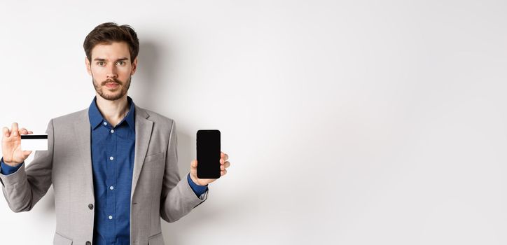 Online shopping. Serious bearded man in business suit showing plastic credit card with empty smartphone screen, standing against white background.