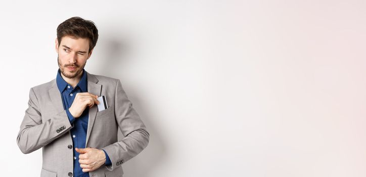 Confident and successful businessman winking aside while place credit card in suit pocket, standing on white background.