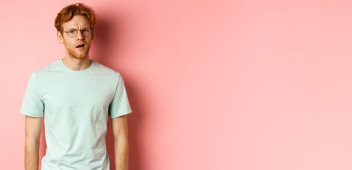 Portrait of handsome redhead man in glasses looking confused, open mouth and stare at camera, cant understand nothing, standing over pink background.