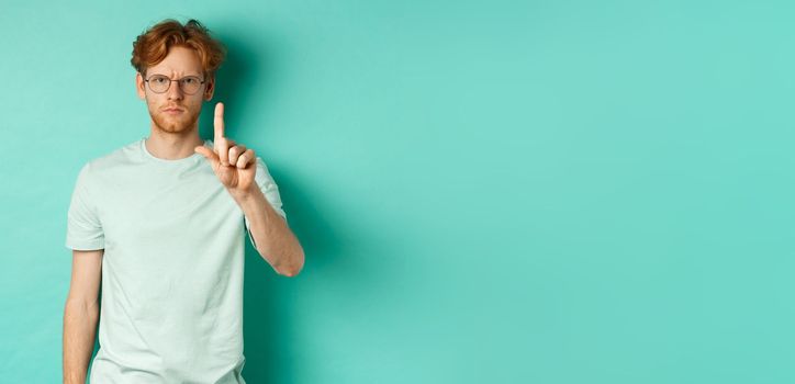 Angry and serious young man with red hair, wearing glasses, showing stop gesture, telling no, shaking finger with disapproval, standing over mint background.