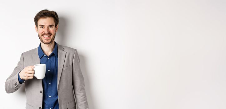 Handsome businessman in suit drinking coffee or tea from office mug, smiling enthusiastic at camera, standing against white background.