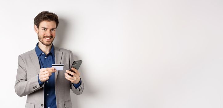 Online shopping. Handsome businessman in suit paying with credit card on smartphone, smiling satisfied at camera, standing on white background.