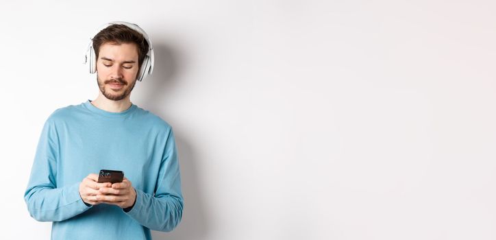 Modern young man in blue sweatshirt put on music on smartphone, listening songs in wireless headphones, white background.