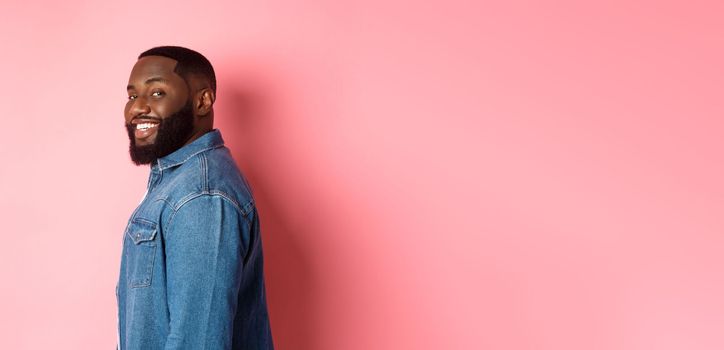 Handsome african-american man with beard, turn face at camera and smiling confident, standing over pink background.