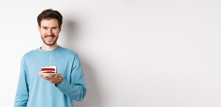 Celebration. Handsome young man celebrating birthday, holding bday cake with lit candle and smiling, making wish, standing over white background.