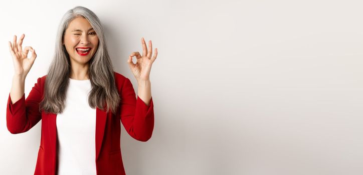 Asian professional businesswoman showing OK signs and winking, smiling pleased, assure or recommend something, standing against white background.