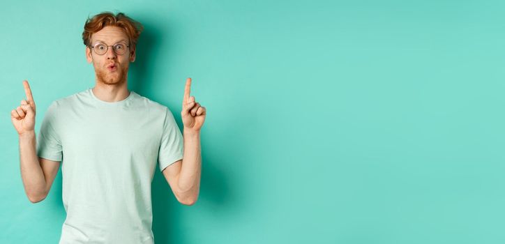 Impressed redhead man in glasses and t-shirt, checking out promo offer, pointing fingers up at copy space, staring at camera amazed, standing over turquoise background.