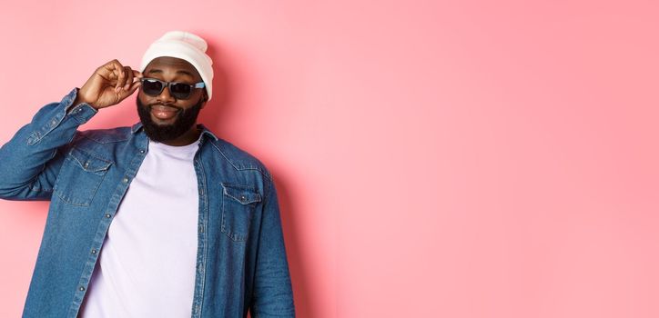 Cool and sassy african-american bearded man, looking confident, touching sunglasses and stare at camera please, standing over pink background.