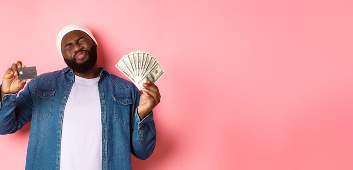 Shopping concept. Sad and whining Black guy showing credit card and dollars, grimacing reluctant, standing over pink background.