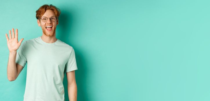 Friendly bearded guy in glasses saying hello, waving hand to greet and welcome you, standing cheerful and smiling over turquoise background.