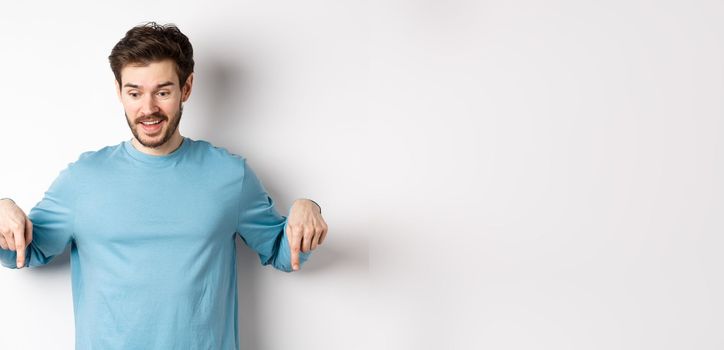 Excited and happy young man looking and pointing down at awesome deal, checking out promotion offer, standing on white background.