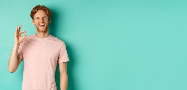 Handsome young bearded man in t-shirt showing Ok sign, smiling with white teeth and saying yes, agree with you, standing over turquoise background.