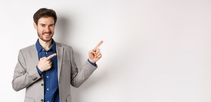 Handsome smiling man in business suit pointing fingers at upper right corner, showing logo, advertising company, standing on white background.