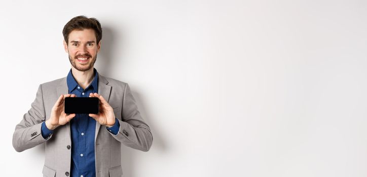 E-commerce and online shopping concept. Happy guy in business suit showing empty smartphone screen horizontally, smiling pleased at camera, white background.