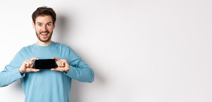 E-commerce and shopping concept. Smiling cheerful man showing empty smartphone screen in horizontal position, looking happy at camera, white background.