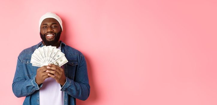 Handsome bearded african-american man showing money, earning dollars, standing over pink background. Copy space