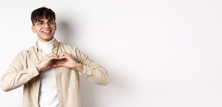 Happy Valentines. Handsome young man in glasses say I love you, showing heart sign and smiling at girlfriend, celebrating lovers day, standing over white background.