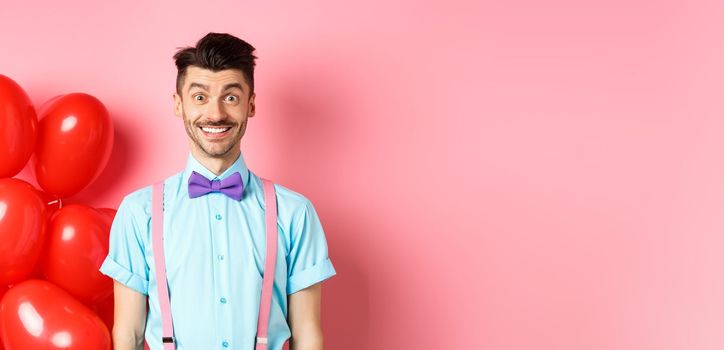 Valentines day concept. Image of handsome young man looking excited and surprised, smiling while standing on romantic pink background near heart balloons.