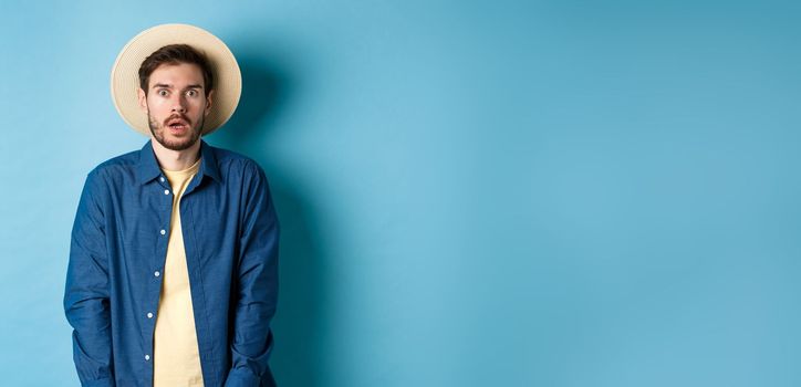 Young man tourist in summer hat drop jaw, staring at something shocking, standing on blue background.