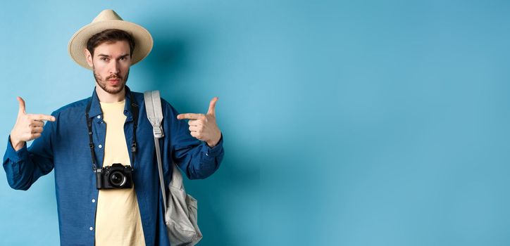 Confident and ready young man pointing at himself, self-promoting or show-off, going on summer vacation, wearing straw hat, holding backpack with camera, blue background.