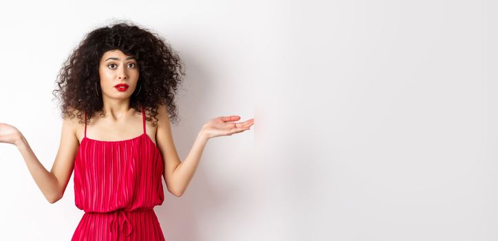 Confused lady in red dress shrugging shoulders, spread hands sideways and looking puzzled, know nothing, cant understand, standing over white background.