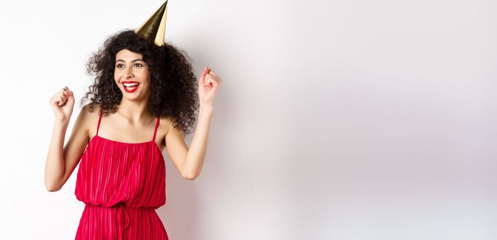 Happy birthday girl celebrating, wearing party hat and red dress, dancing and having fun, standing against white background.
