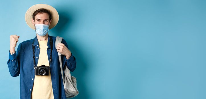 Covid-19 and summer holidays concept. Happy young man travelling abroad in medical mask, saying yes and raising fist up joyfully, cheerful about vacation, blue background.