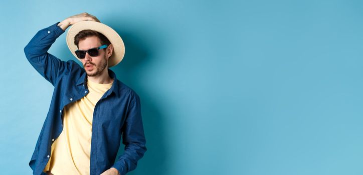 Cool guy having fun on vacation, wearing straw hat and sunglasses, looking aside sassy, standing on blue background.