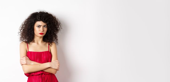 Sad and offended caucasian woman in red dress frowning, cross arms on chest and sulking, feeling mad at you, standing over white background.