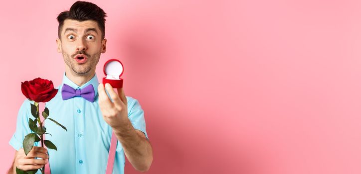 Valentines day. Funny man with moustache and bow-tie making proposal, showing engagement ring and propose with red rose, standing over pink background.