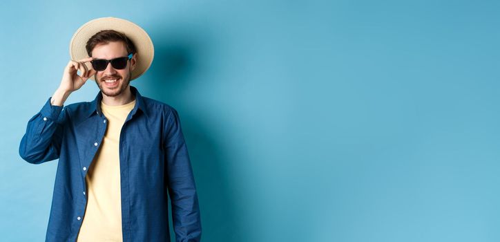 Happy smiling guy going on summer vacation, wearing straw hat and black sunglasses, standing on blue background.