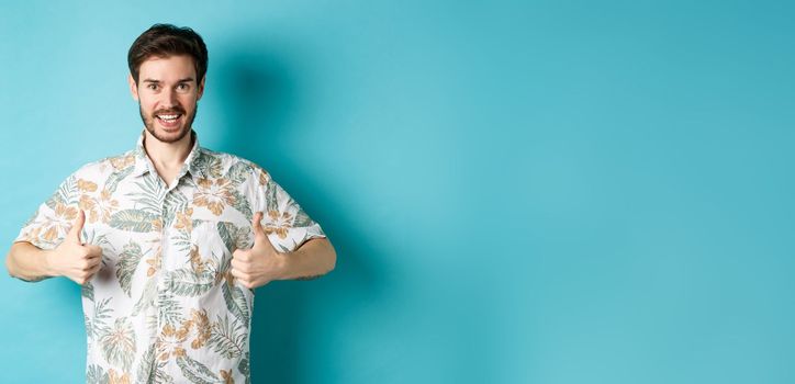 Handsome tourist showing thumbs up and say yes, praising good tourism agency, wearing hawaiian shirt, standing on blue background.