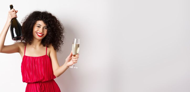 Happy woman partying on valentines day holiday, dancing with glass and bottle of champagne, wearing red dress, smiling on white background.