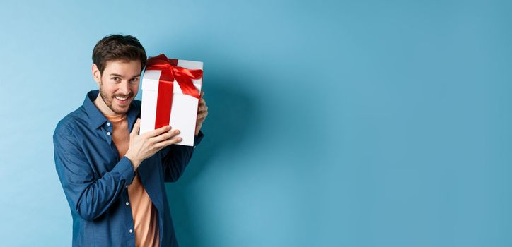 Valentines day. Romantic boyfriend showing gift box for lover, celebrating anniversary, standing over blue background.