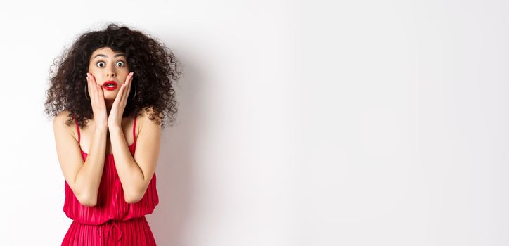 Shocked and startled woman staring at something amazing, touching face and looking at camera, wearing red dress on white background.