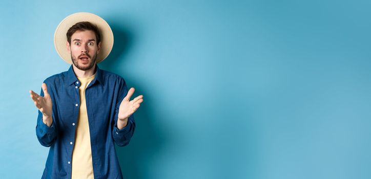 Shocked male tourist on vacation raising hands up and looking at camera startled, react to big news with disbelief, standing on blue background in straw summer hat and shirt.