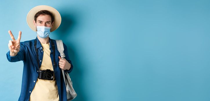 Covid-19, pandemic and travel concept. Positive guy going on vacation, wearing summer hat and medical mask, showing peace sign, standing with backpack and camera on blue background.