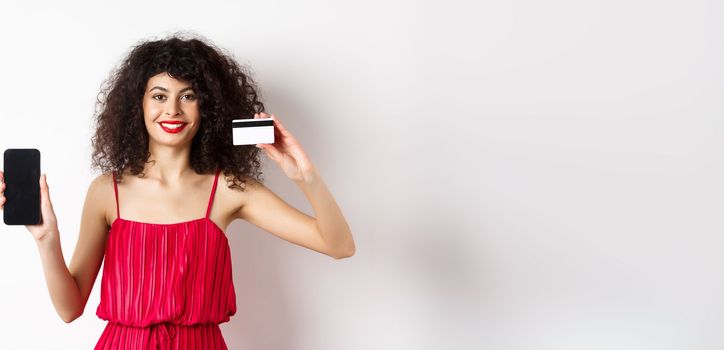 Online shopping concept. Elegant female model with curly hair, wearing red dress, showing plastic credit card with empty mobile phone screen, standing on white background.