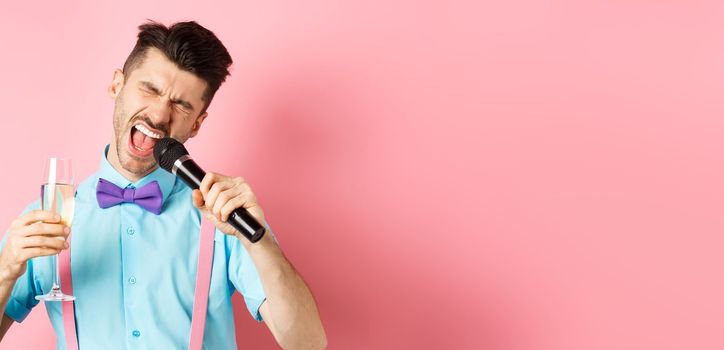 Party and festive events concept. Drunk funny guy singing in microphone and drinking champagne from glass, having fun at karaoke bar, standing on pink background.